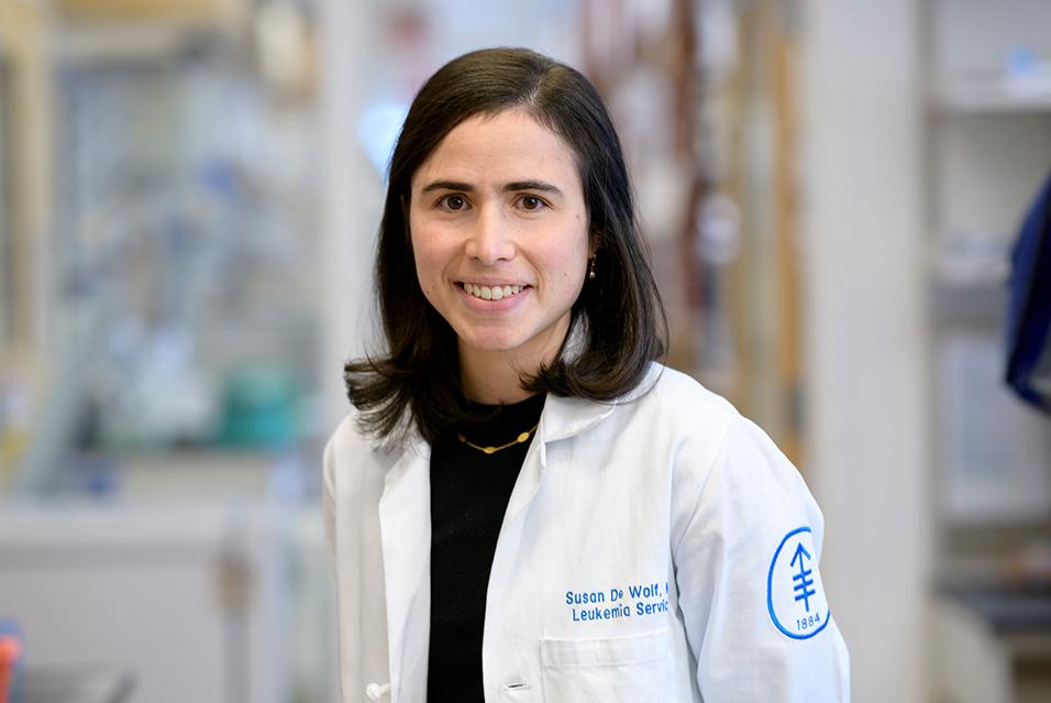 Woman with shoulder-length dark brown hair wearing a white lab coat with the MSK logo on it, standing in an MSK lab and smiling 