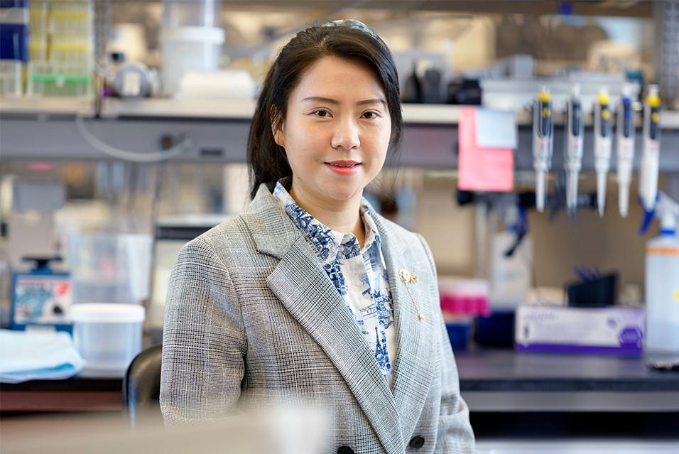 Woman with her hair pulled back, wearing a gray blazer and white and blue shirt, standing in a lab and smiling 