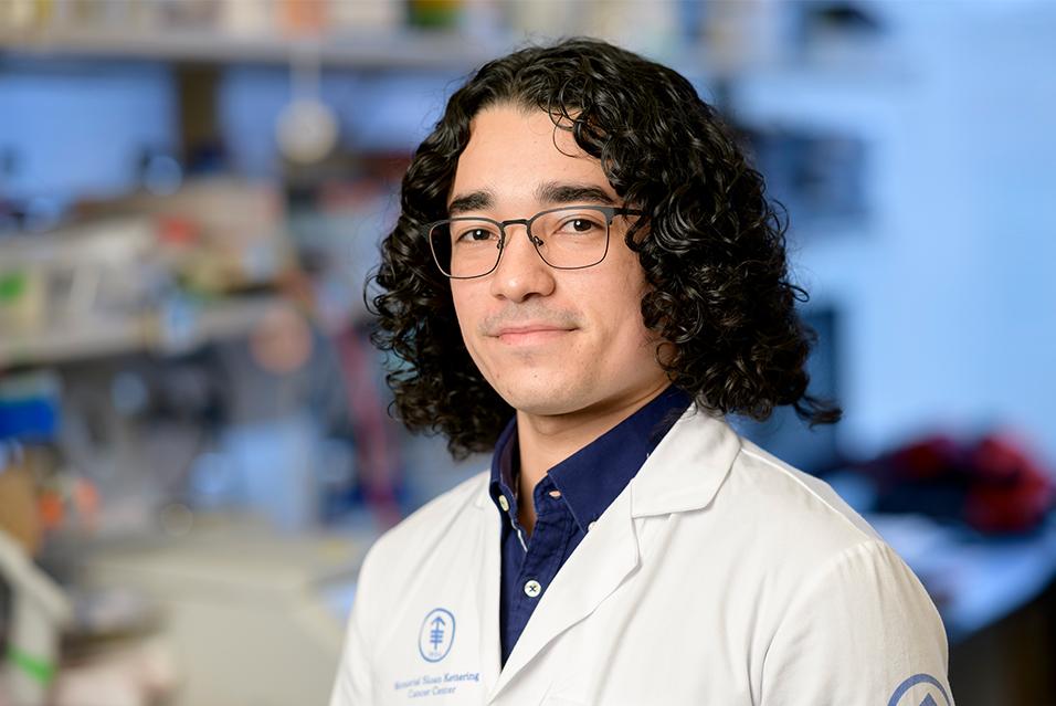 Man with dark, chin-length curly hair wearing glasses and a white lab coat with the MSK logo and a blue collared shirt, smiling 