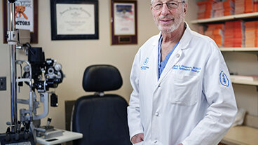 Doctor in a white coat standing in an exam room with ophthalmology equipment.